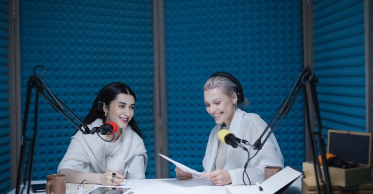 Duas mulheres sorrindo em estúdio de gravação de rádio com microfones vermelhos e amarelos.