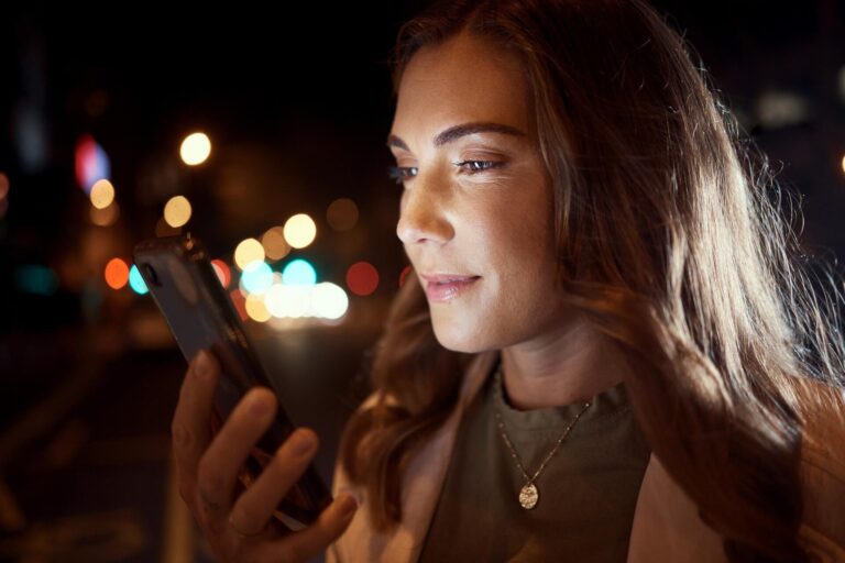 Anúncios na Black Friday ficam 45% mais caros no Facebook e no Instagram Mulher de cabelos longos olha a tela do smartphone à noite, com luzes desfocadas da cidade ao fundo
